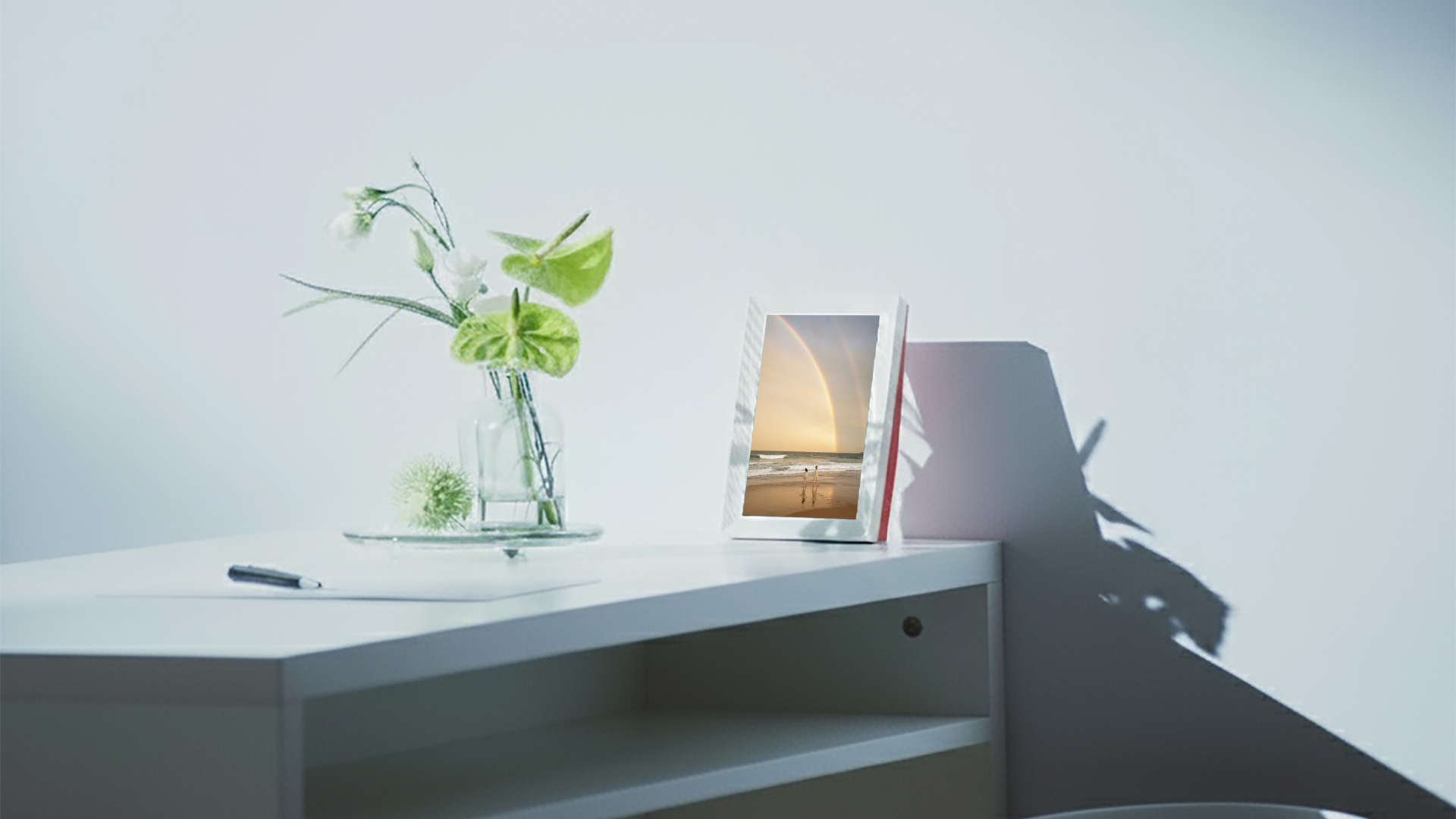 Digital photo frame on a white surface with a vase of flowers and a pen in the background.